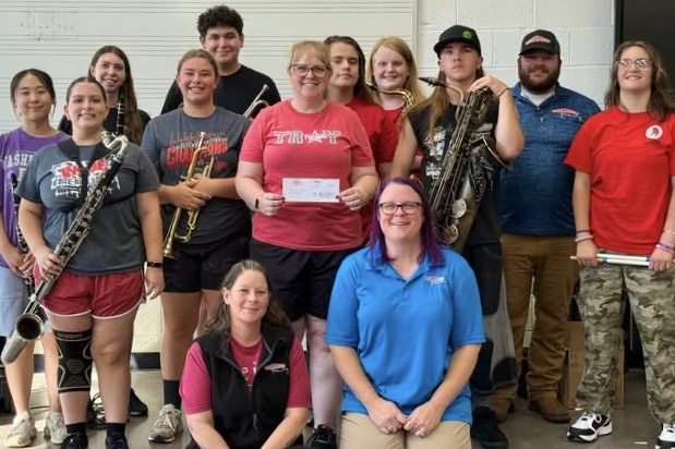 A group of high school band members stand together smiling, some holding their instruments, while a woman at center holds up a check.