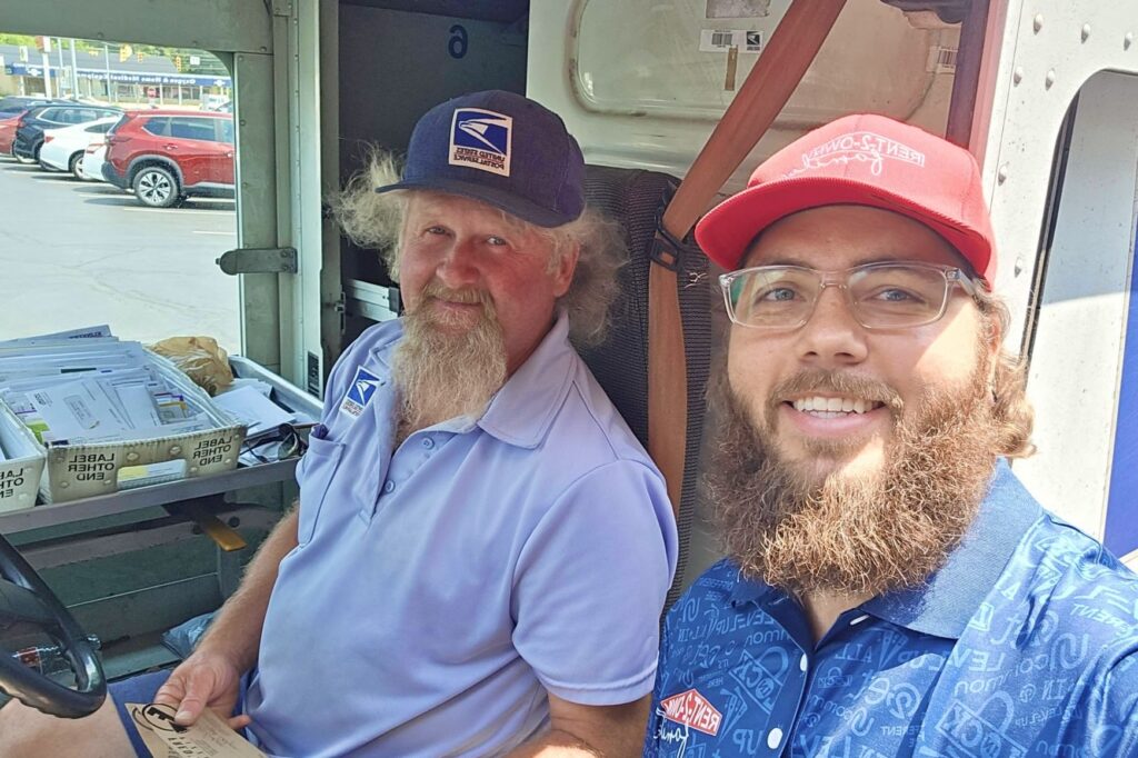 Two men smile; one is wearing a USPS uniform, sitting in a postal truck, and holding two gift certificates.