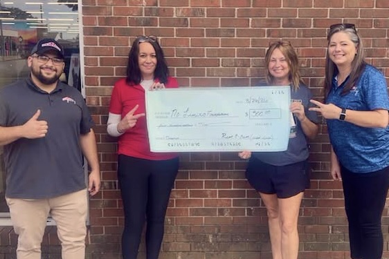 Four people stand smiling in front of a brick building; two of them hold up an oversized check for $500 from R2O to the No Limits Foundation.