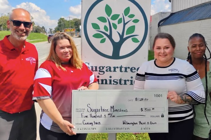Four people stand smiling in front of a Sugartree Ministries sign; two hold up an oversized check for $500 from R2O to Sugartree Ministries.