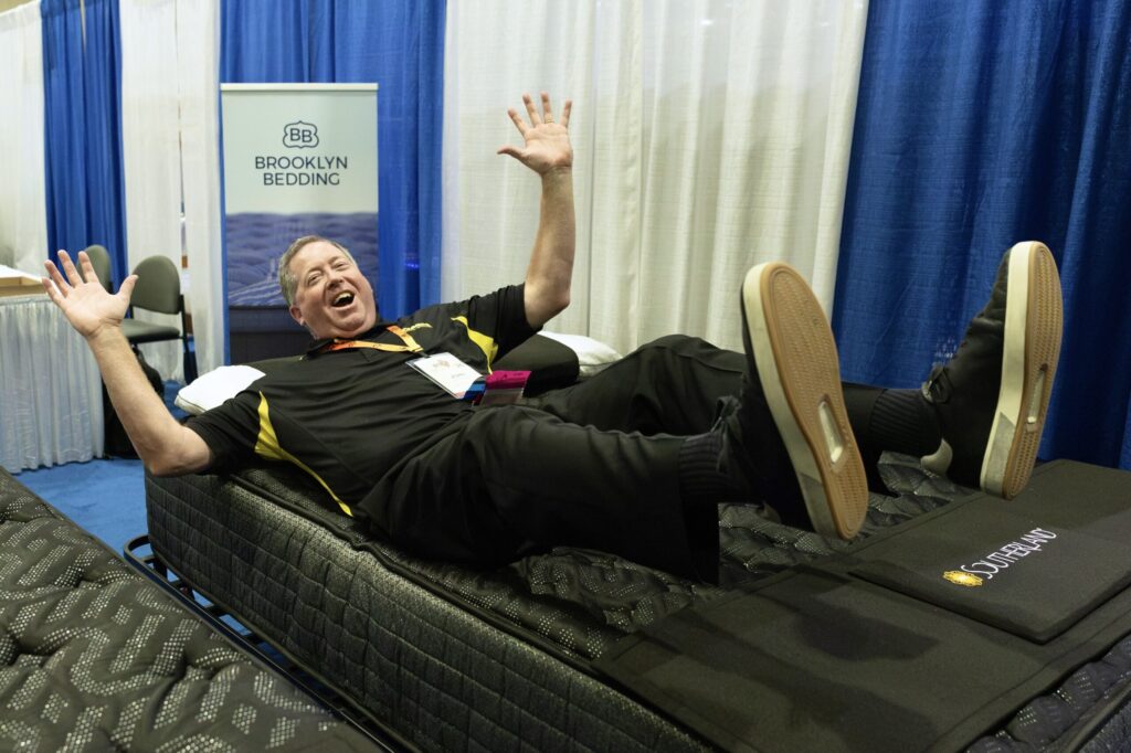 A laughing man falls backward onto a mattress next to a sign reading “Brooklyn Bedding.”