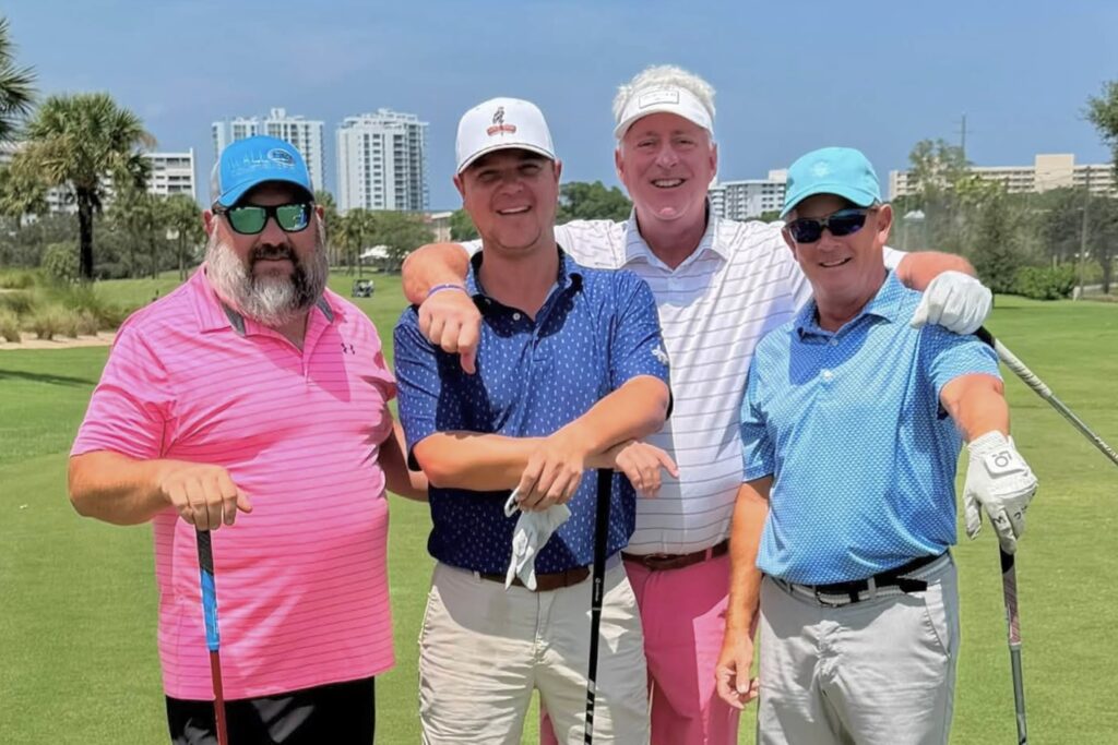 Four vibrantly clad men holding golf clubs and wearing caps cluster together smiling on a bright green golf course.