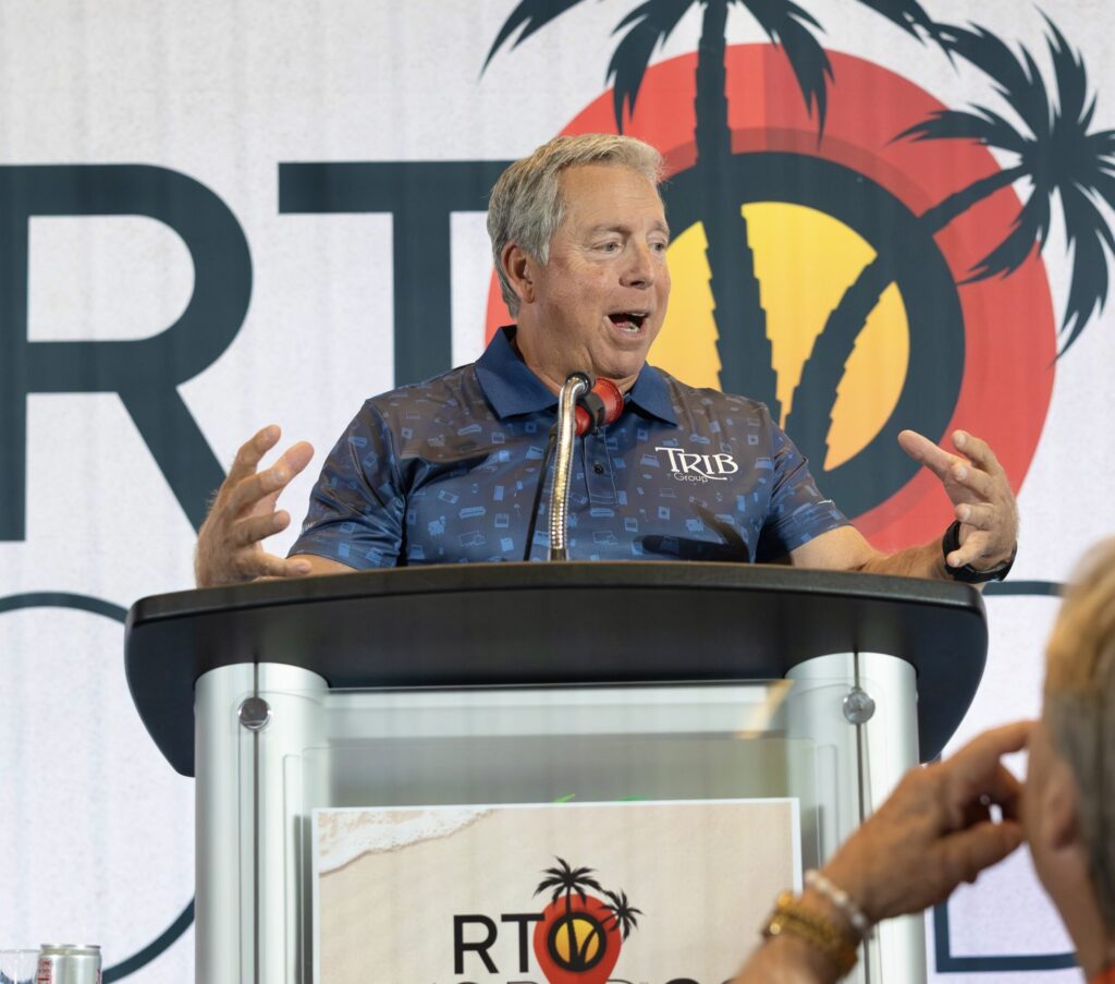 An expressive man stands behind a podium reading “RTO World 26,” arms outstretched and mouth agape.