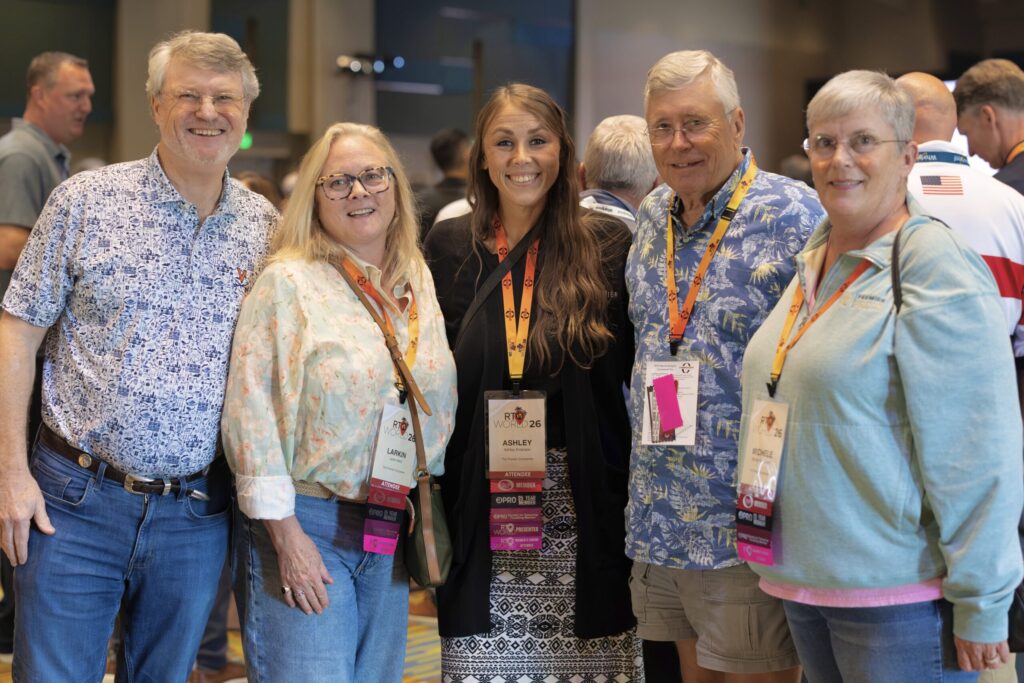 Five people wearing lanyards and nametags stand together smiling with a crowd behind them.