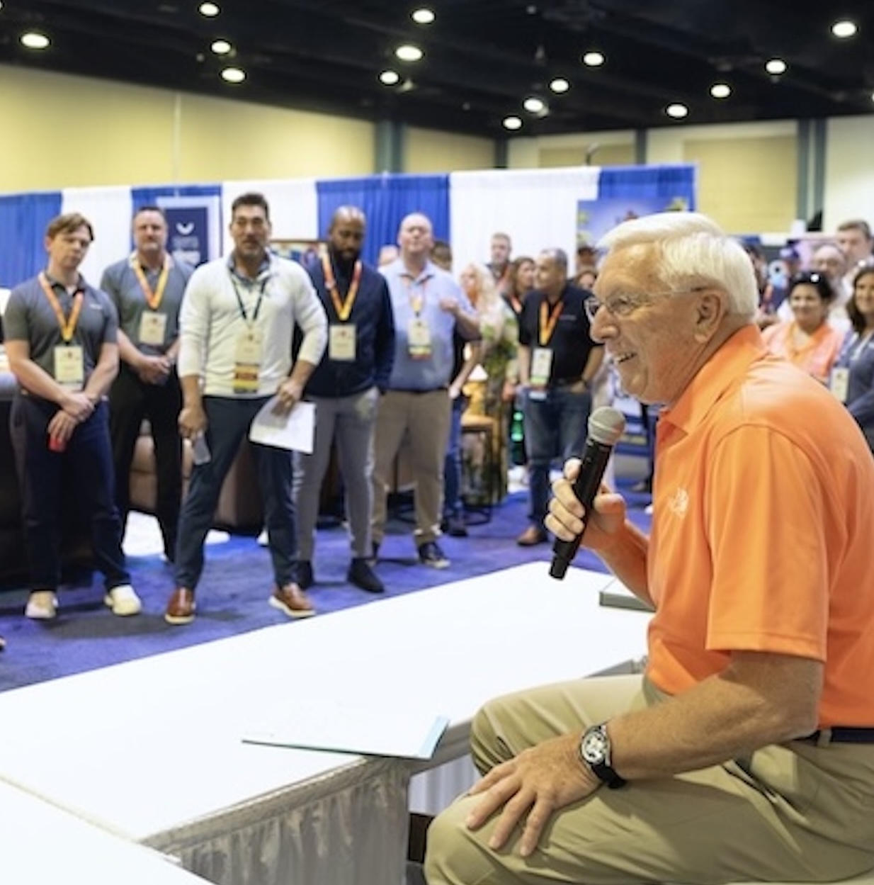 A man with white hair and an orange shirt sits speaking into a microphone as he faces a small standing group of people.
