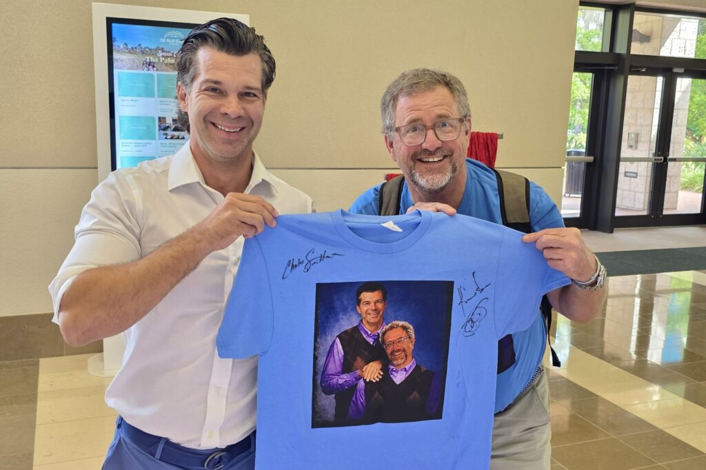 Two men smile as they hold up a bright blue t-shirt with a painted-style illustration of the two of them, along with two signatures, on it.