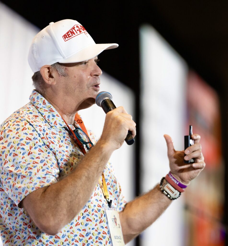A man wearing a baseball cap and a colorful shirt animatedly speaks into a microphone.
