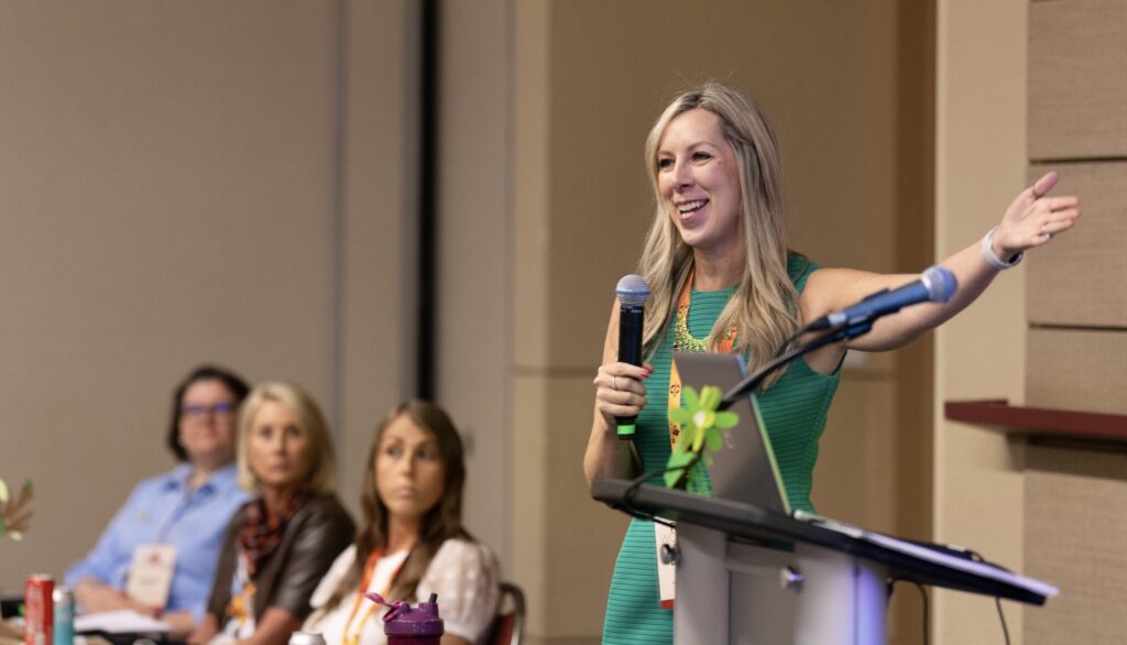 A blonde woman holds a microphone in one hand, with her other arm outstretched, as she energetically addresses a panel and an audience of women.