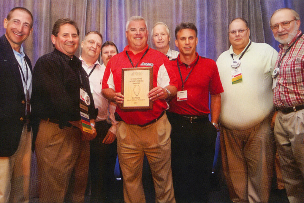 A group of men stand together smiling as one man holds up a gold and wooden plaque.