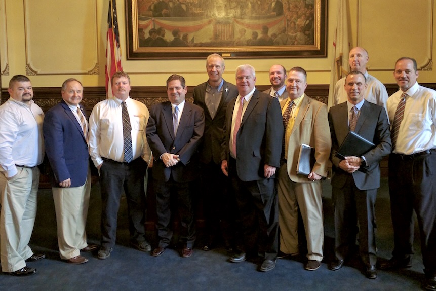 A group of men wearing suits stand together smiling with flags and a large historic painting behind them.
