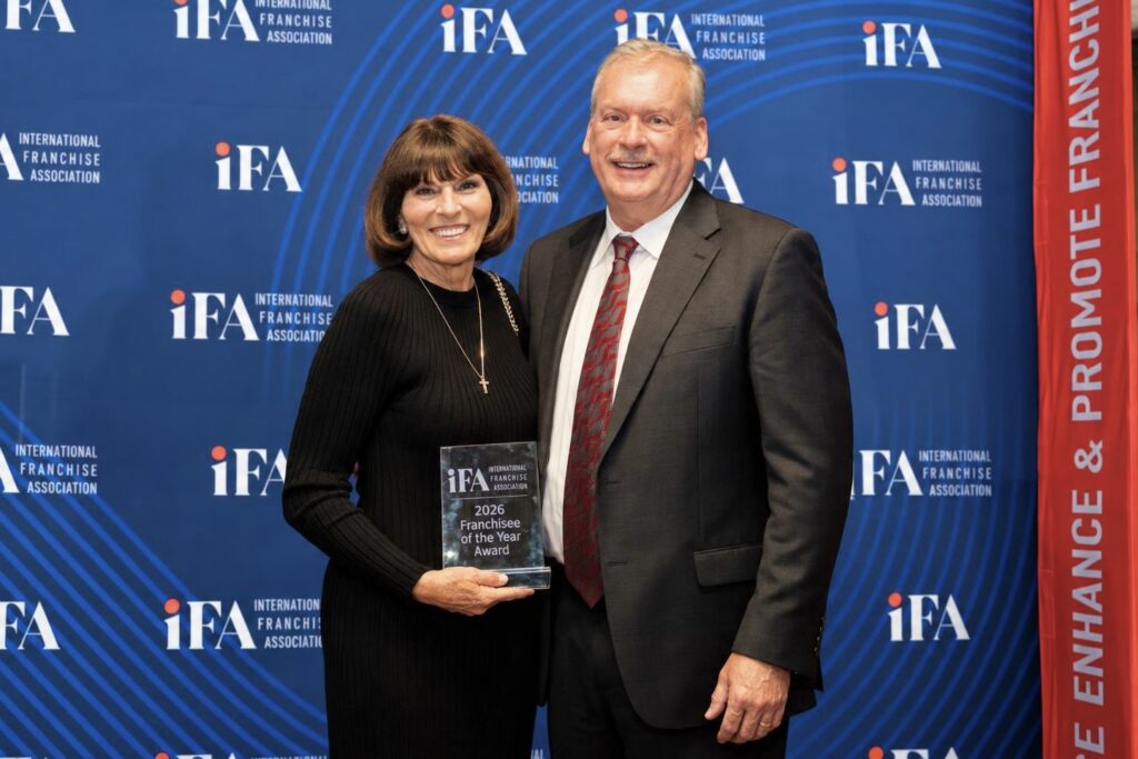 A woman and man wearing cocktail attire stand together holding a plaque and smiling. Behind them is a blue wall with the International Franchising Association logo all over it.