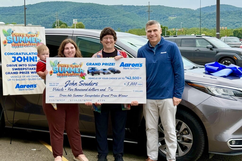 A man and a woman holding a baby smile and hold up an oversized check for $5,000 from Aaron’s to John Souders, as another man looks on smiling. Behind them is a silver minivan with a huge blue bow stuck on its hood, and a sign reading “Congratulations, John! Drive Into Summer Sweepstakes Grand Prize Winner.”