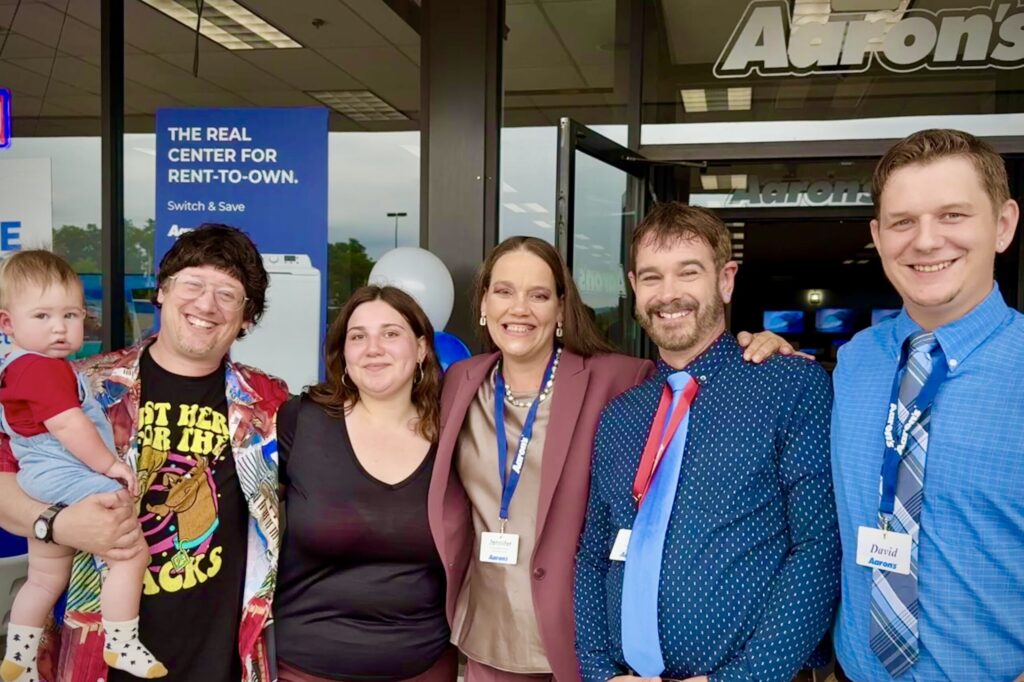 A group of five people stand together at an Aaron’s store entrance smiling. Three of them are wearing Aaron’s lanyards and nametags.