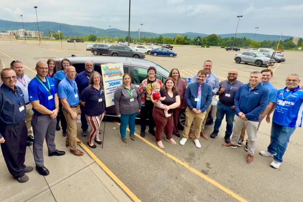 A large group of people stand together in a large parking lot smiling. Behind them is a silver minivan with a huge blue bow stuck on its hood, and a sign reading “Congratulations, John! Drive Into Summer Sweepstakes Grand Prize Winner.”