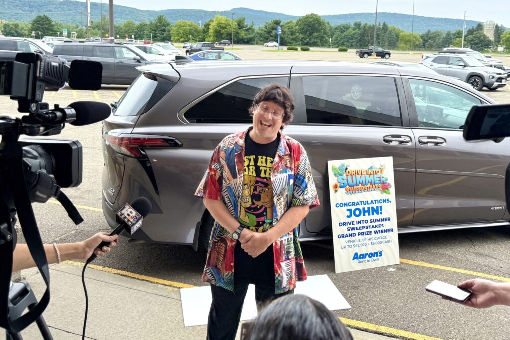 A man wearing a bright tee and aloha shirt stands in a parking lot and animatedly talks toward a gathering of cameras and microphones. Behind him is a silver minivan and a sign reading “Congratulations, John! Drive Into Summer Sweepstakes Grand Prize Winner.”
