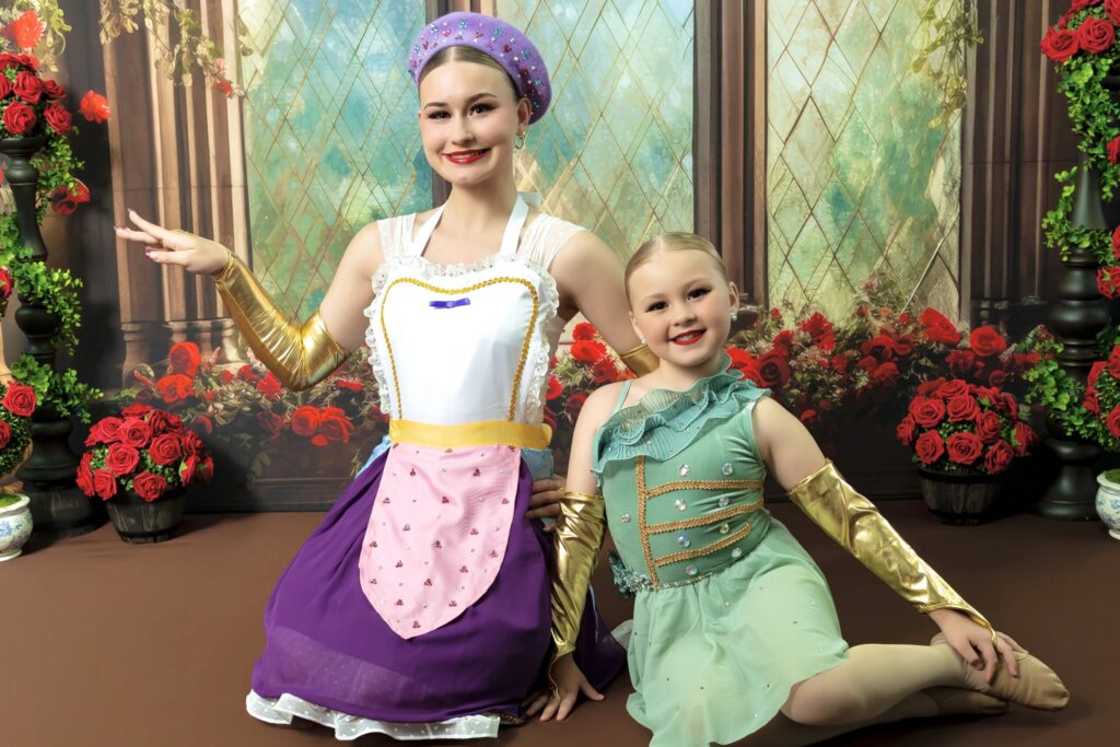 A young woman and a young girl, both in full stage makeup and colorful, glittery costumes, smile and pose on a stage with a painted backdrop behind them.