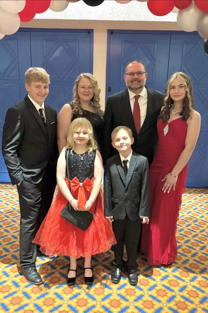 A family of six – a father, mother, older boy, older girl, younger boy, and young girl – stand together smiling. All are wearing semi-formal attire and stand beneath a black, white, and red balloon arch.