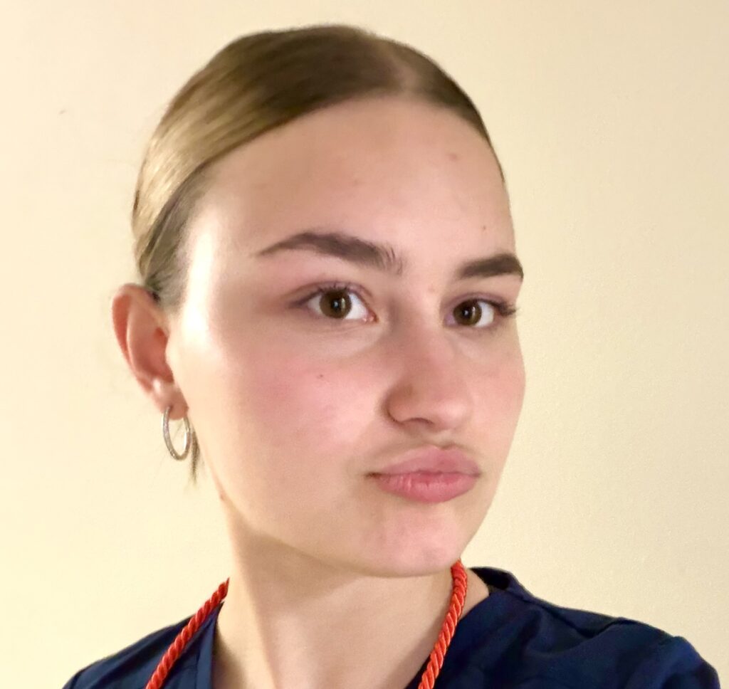 A young blonde woman wearing blue scrubs and a maroon cord draped around her neck poses with ‘duck lips.’