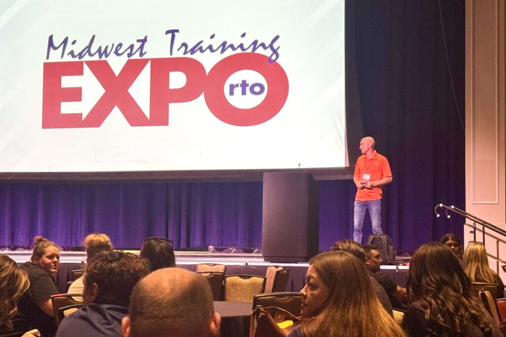 A man wearing a bright shirt and blue jeans holds a microphone while standing up on a stage in front of a crowd, with a giant projection of the Midwest RTO Training Expo logo behind him.