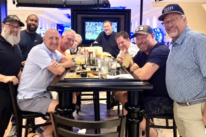 A group of men sit and stand around a long table full of dishes and glasses, all smiling.