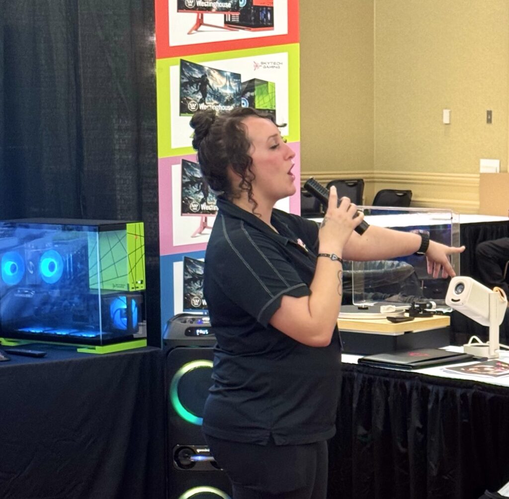 A young woman wearing all black stands by a few tables full of products, singing into a microphone.