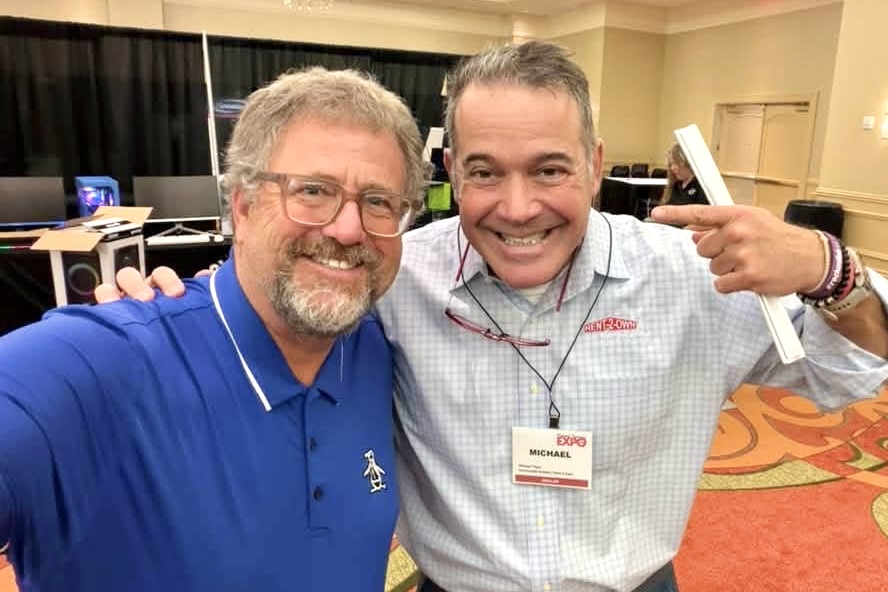Two men stand with their faces close to each other, smiling broadly.