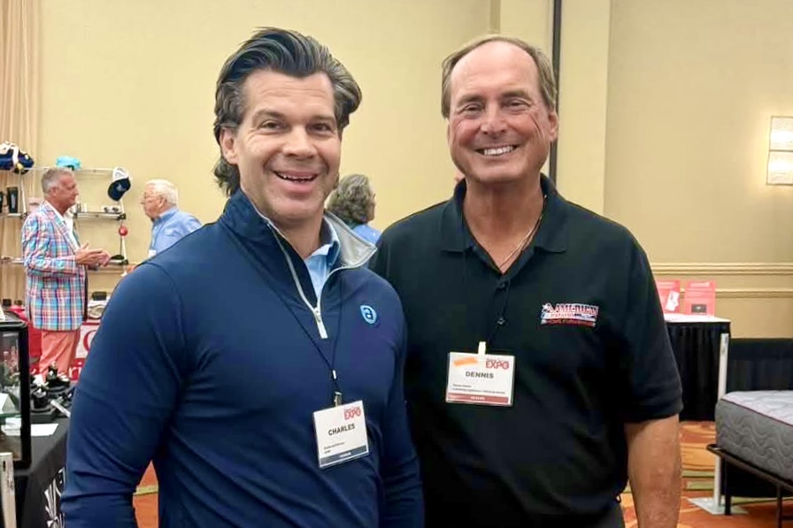 Two men wearing logo shirts and nametags stand together smiling, as others converse behind them.