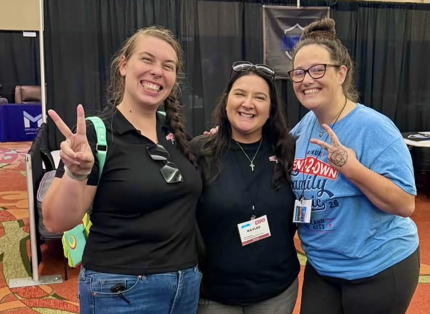 Three women stand with their arms around each other, two of them showing ‘peace’ fingers, and smiling, with a table full of jewelry displayed in front of them.
